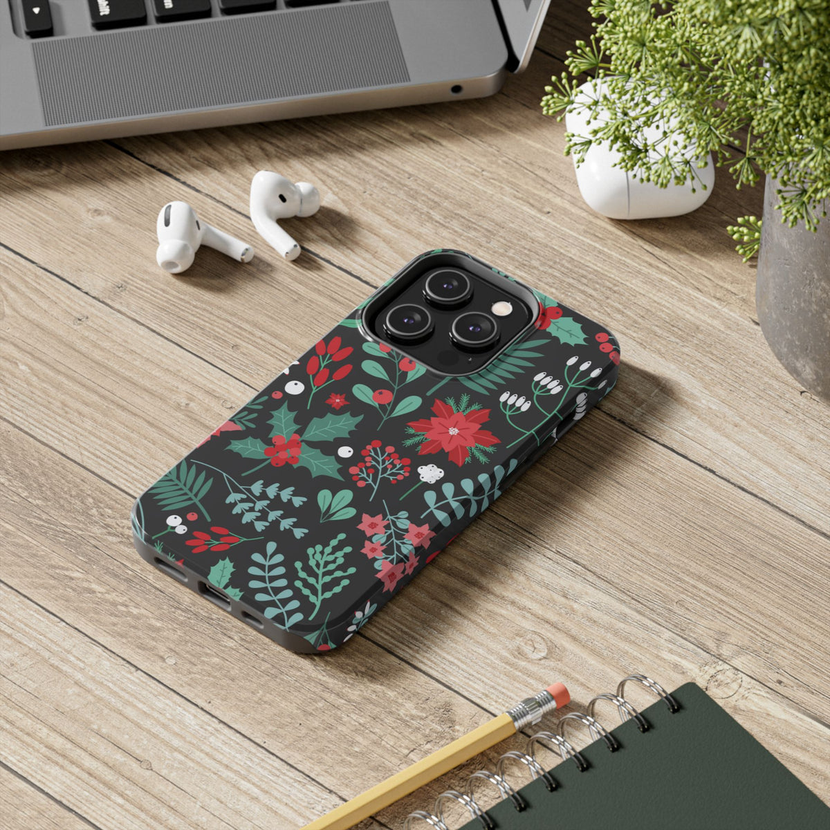 Midnight Poinsettia tough phone case on a wooden desk next to AirPods and a notebook