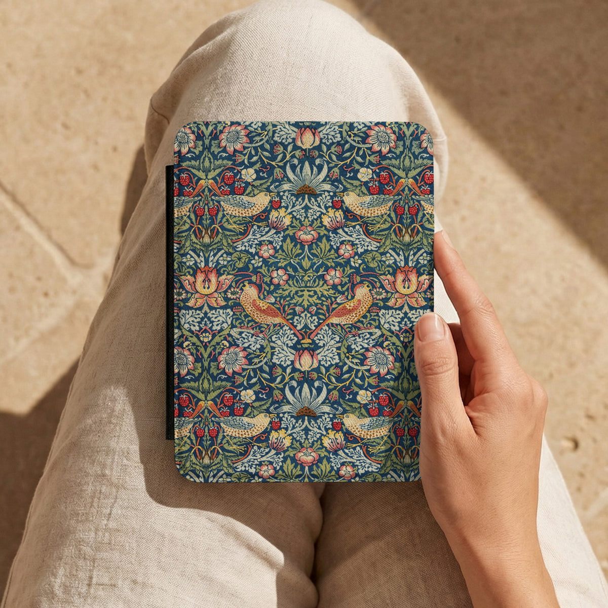 Hand holding a Morris Strawberry Thief Kindle case on a lap