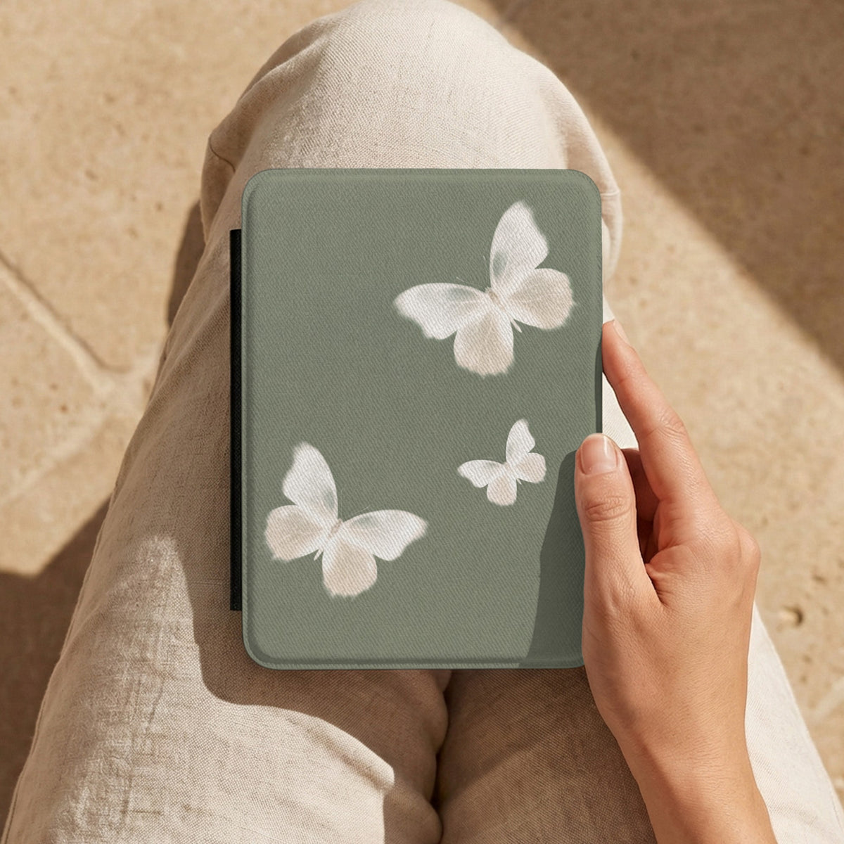 Sage Butterfly Kindle case held on a lap in linen trousers